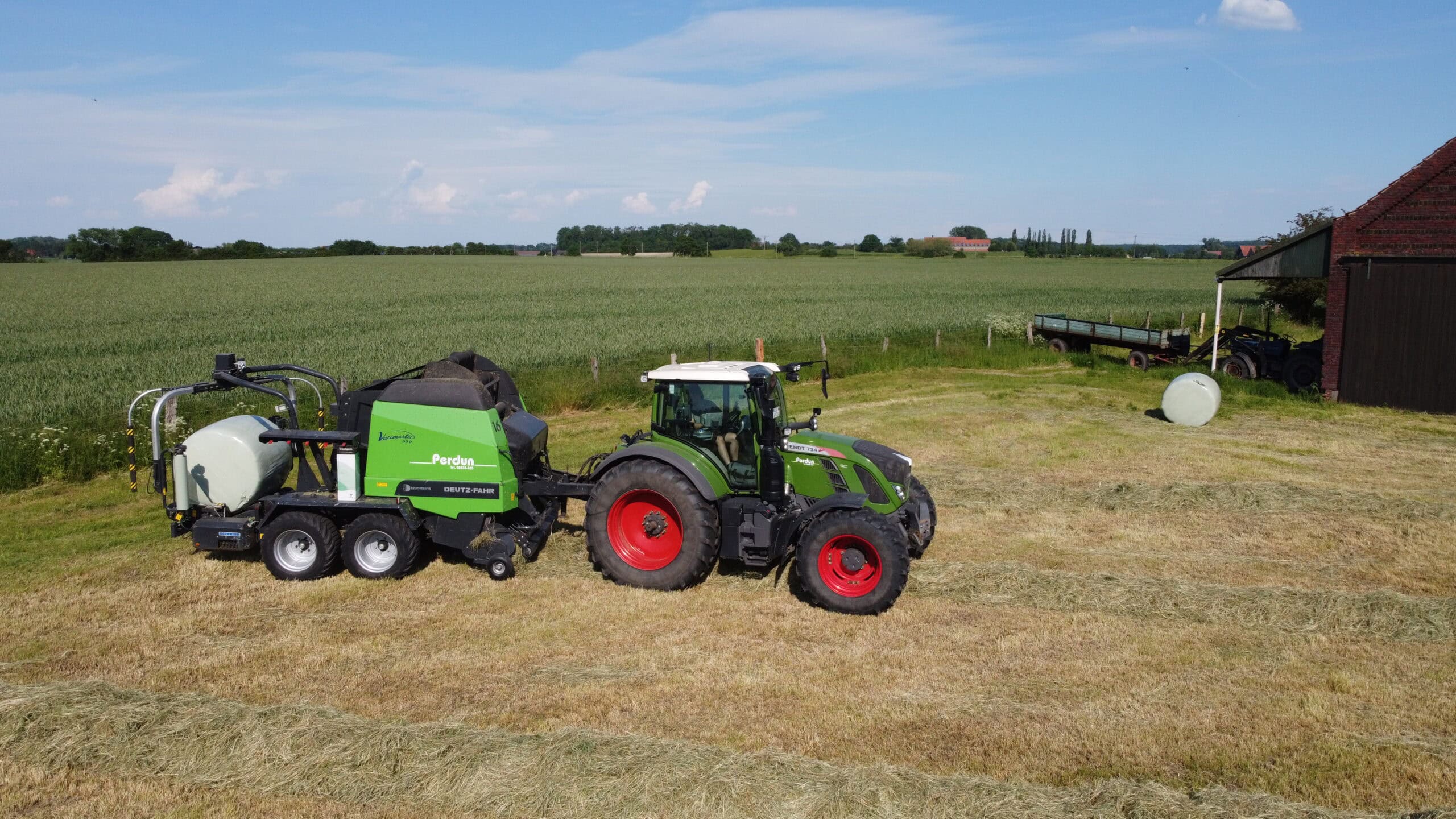 Ein grüner Traktor mit rotem Siloanhänger arbeitet auf einem Heufeld neben einer Scheune.