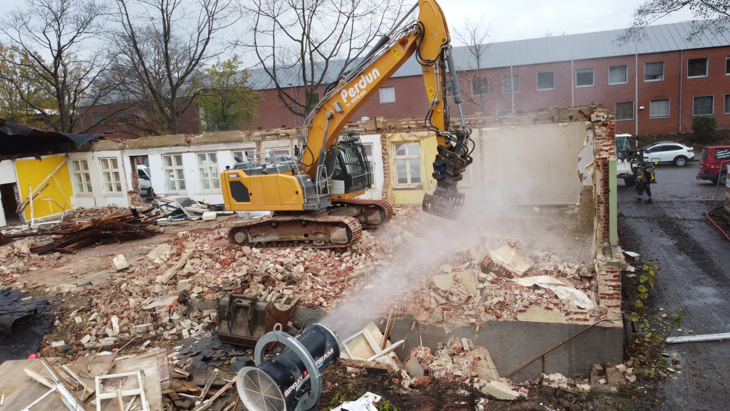 Ein gelber Bagger reißt ein Gebäude ab; herumliegende Trümmer und Sprühwasser auf der Baustelle.