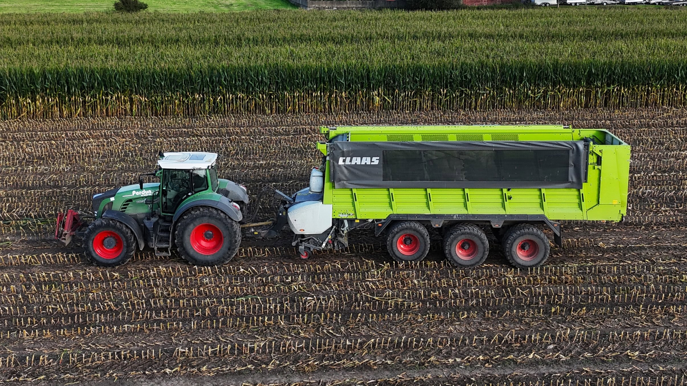 Ein grüner Traktor mit einer grünen Claas-Anhängermaschine auf einem abgeernteten Feld, Vogelperspektive.