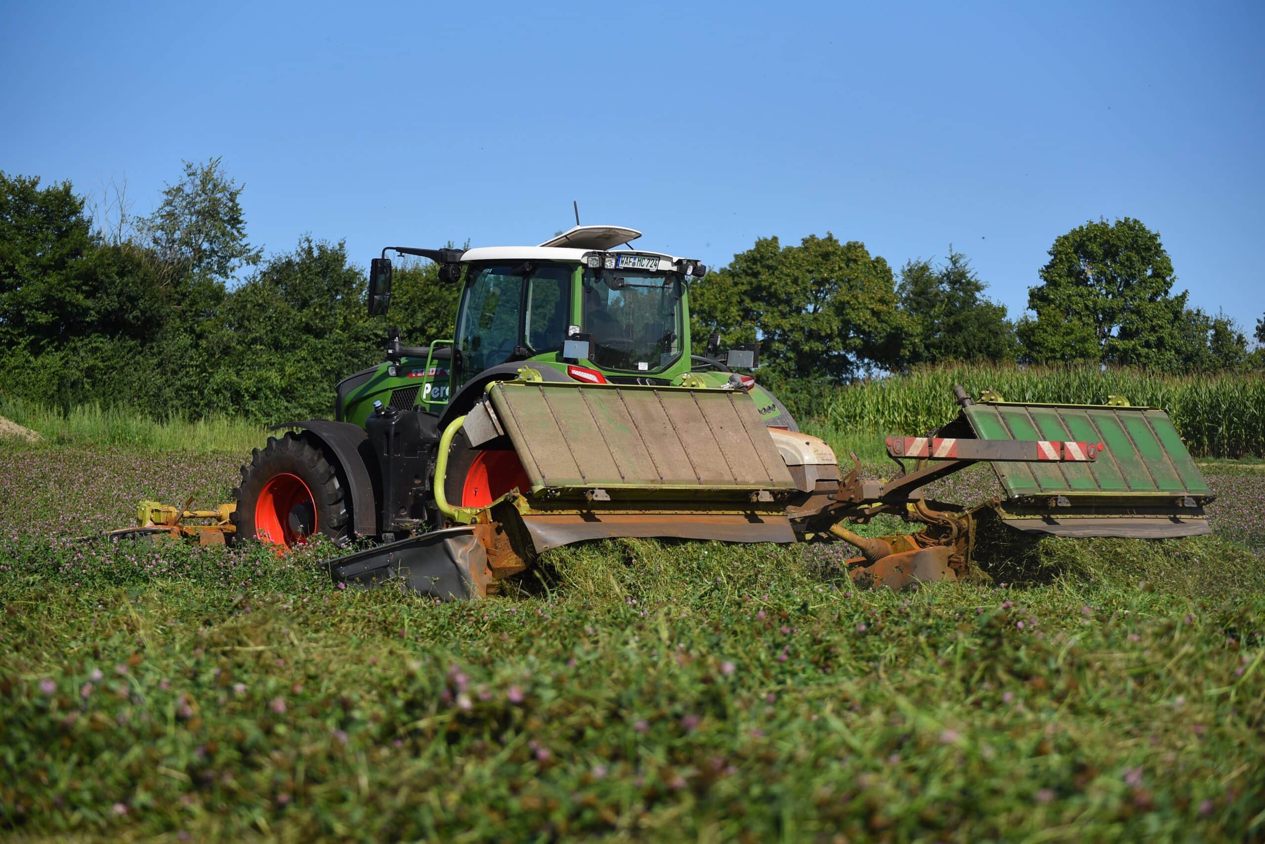 Ein grüner Traktor mäht Gras auf einem Feld unter klarem, blauem Himmel mit Bäumen im Hintergrund.