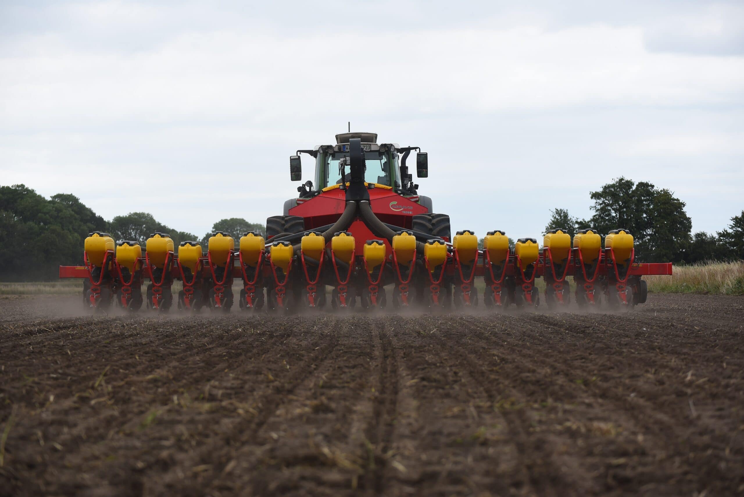 Ein roter Traktor mit gelber Anhängesämaschine fährt auf einem braunen Feld bei bewölktem Himmel.