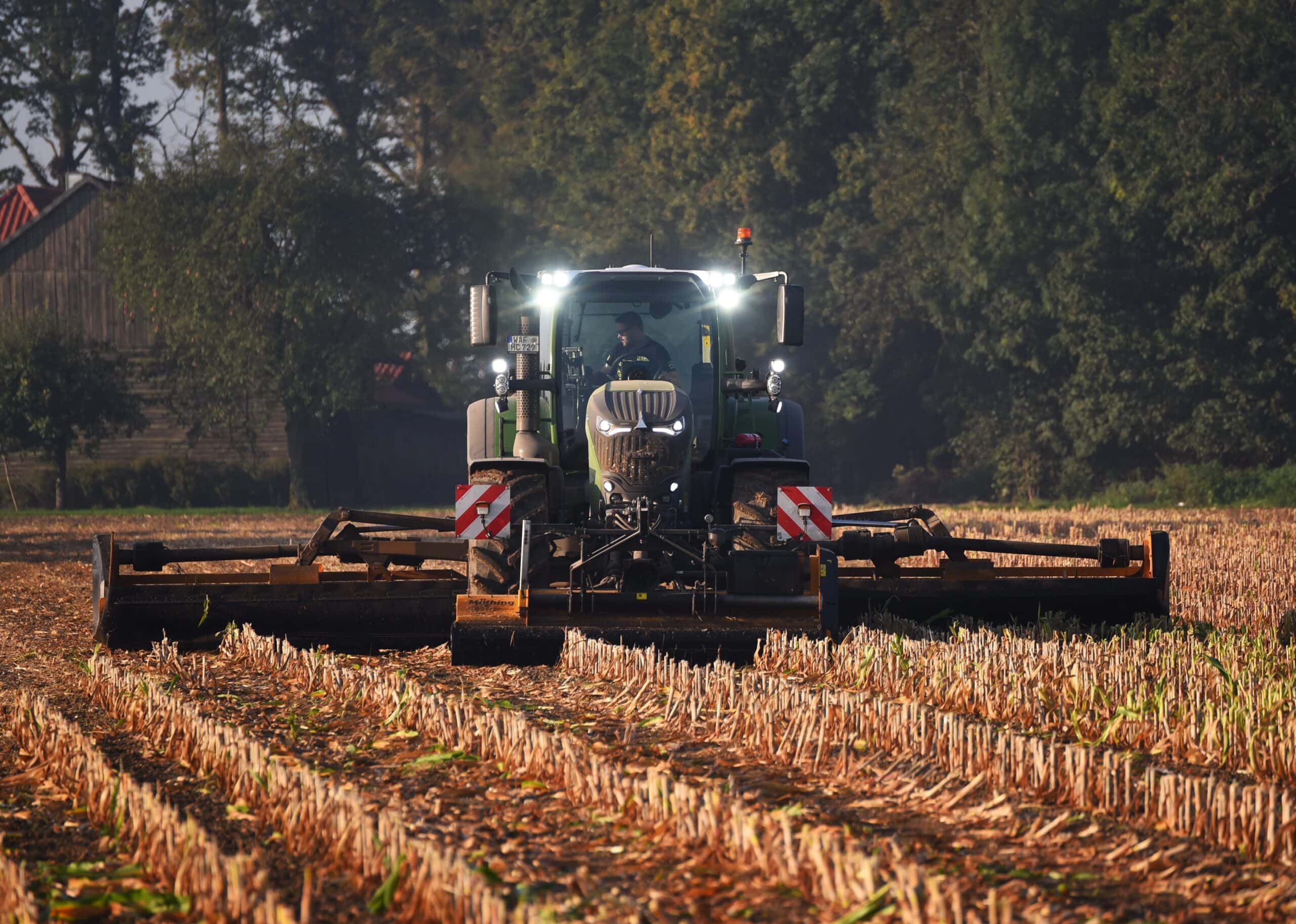 Traktor pflügt Feld, Lenker nicht sichtbar, hellgrün, helle Scheinwerfer, dichte Bäume im Hintergrund.