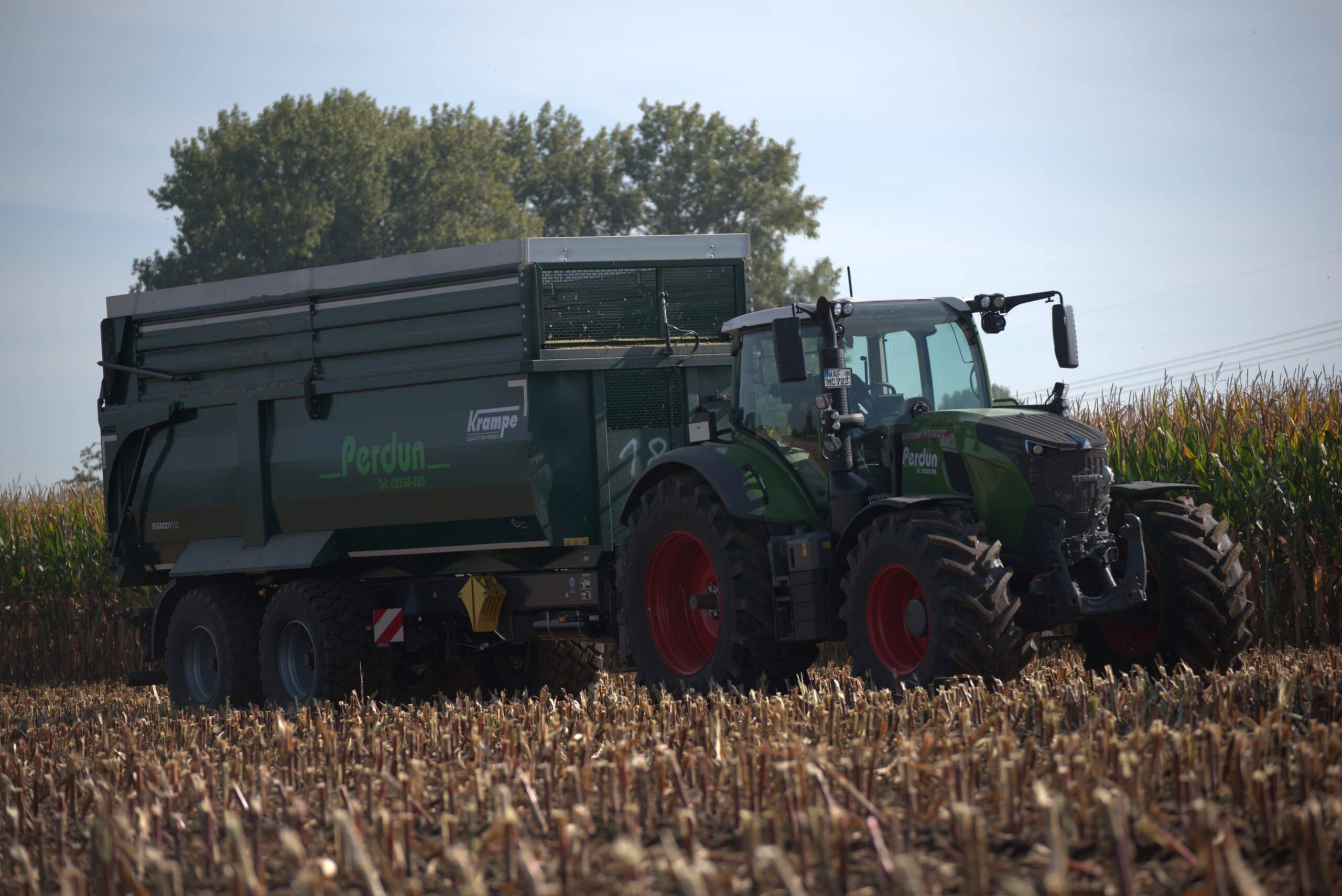 Ein grüner Traktor mit Anhänger auf einem Maisfeld, dunkelgrüne Farbe, Herbsternte, De_DE_formal.