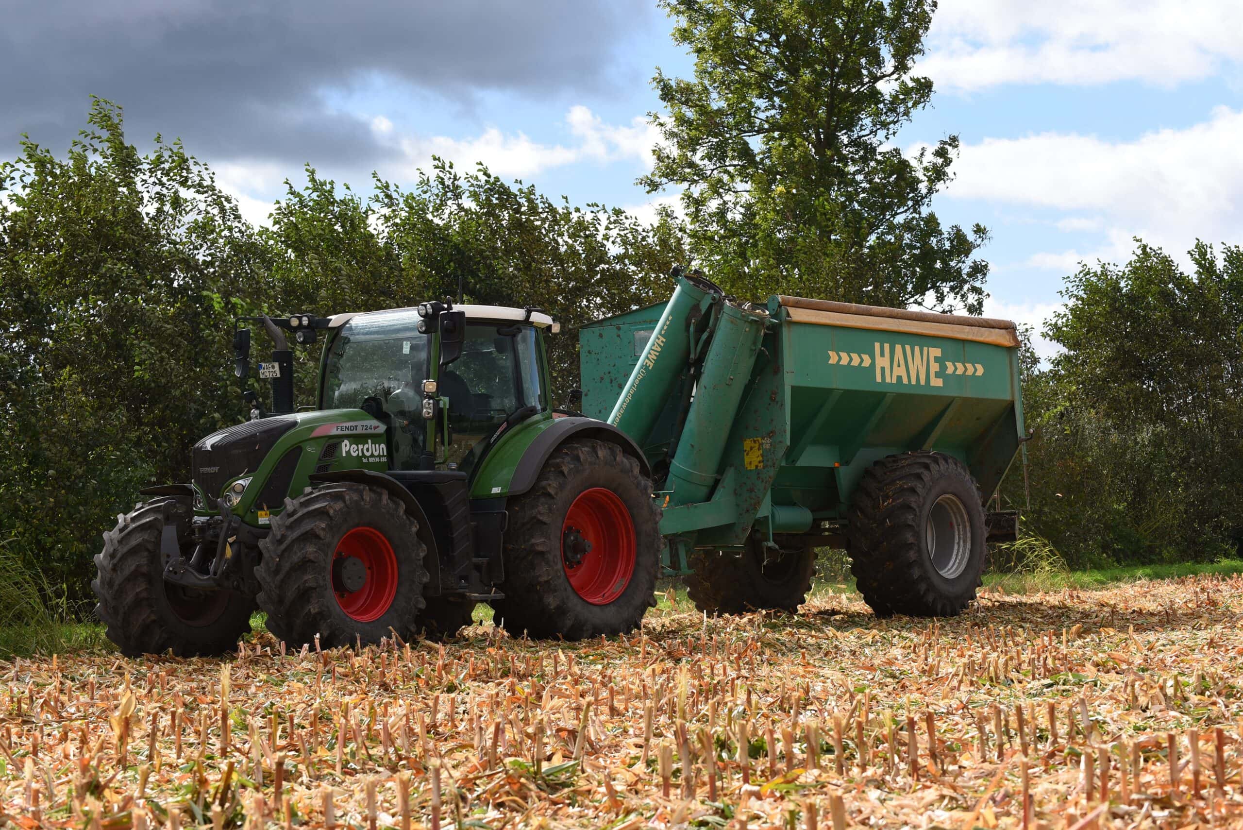 Traktor auf Feld mit angehängtem HAWE-Anhänger, Wolken am Himmel, Bäume im Hintergrund.