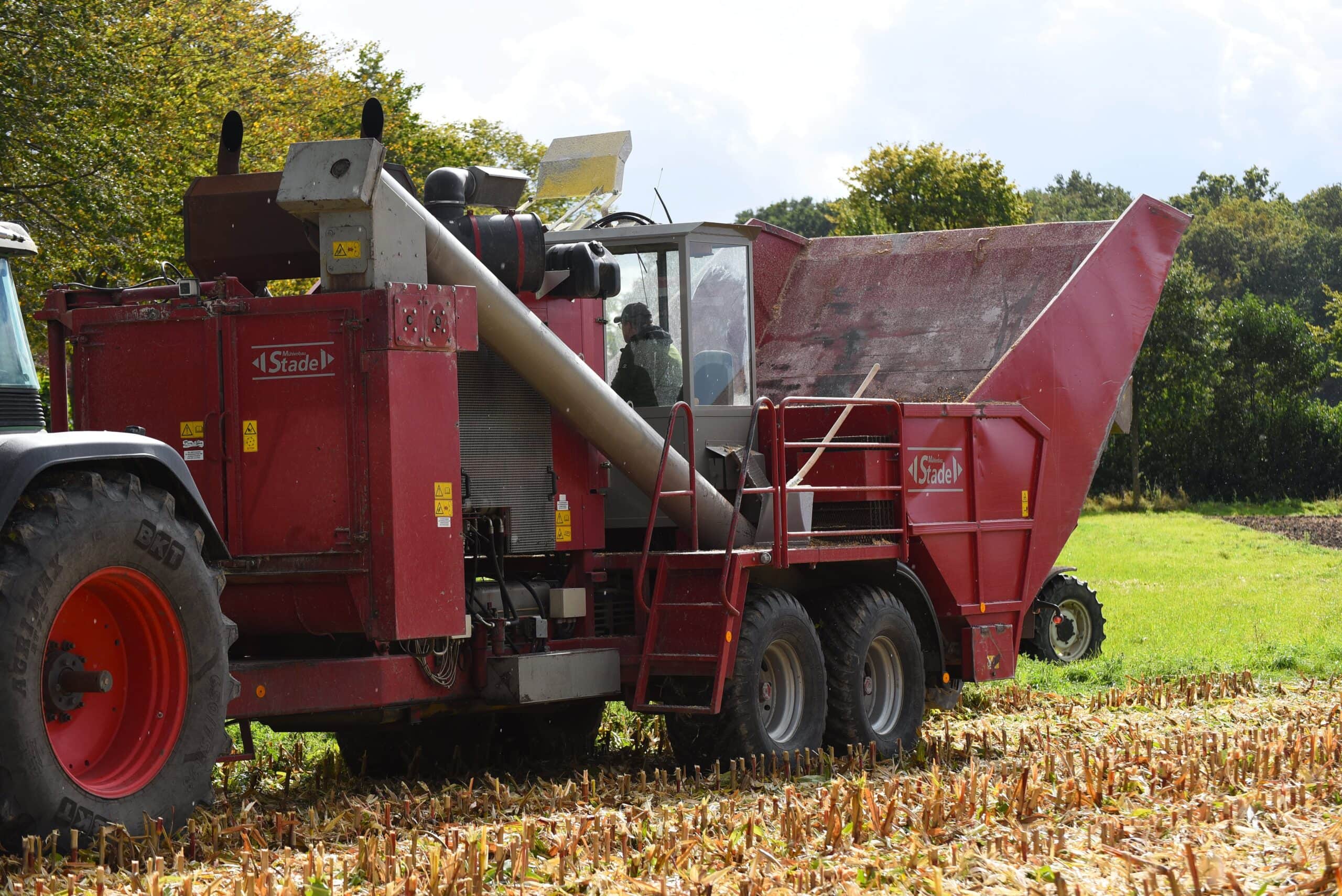 Eine rote Erntemaschine in einem Feld bei der Arbeit. Ein Traktor links, grüne Landschaft im Hintergrund.