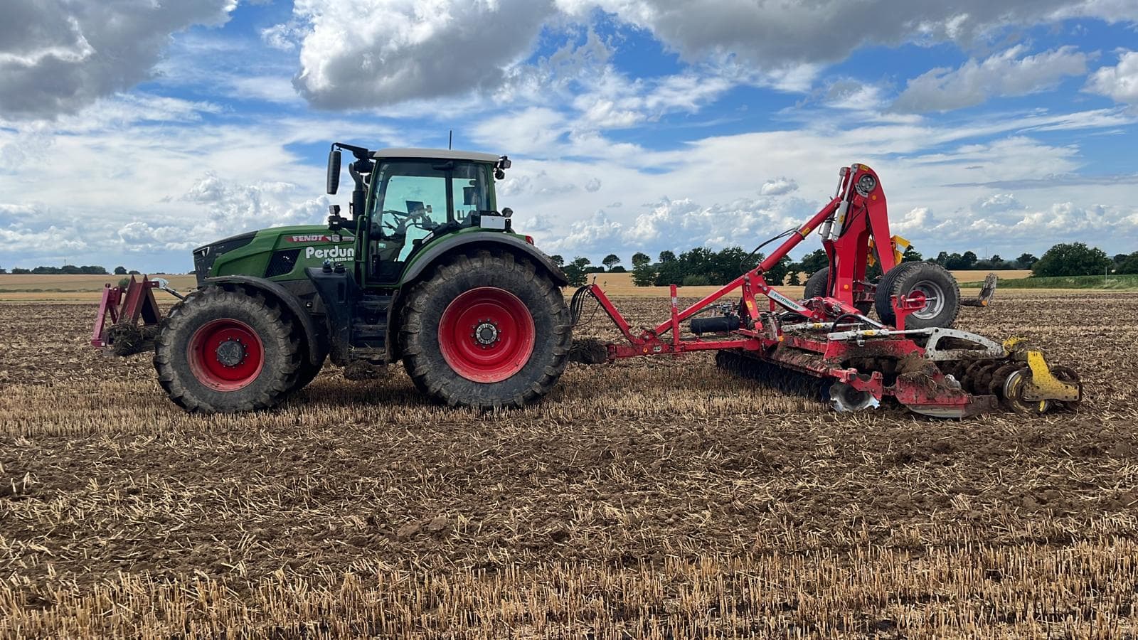 Traktor auf einem Feld, grün und rot, mit landwirtschaftlichem Gerät unter bewölktem Himmel.