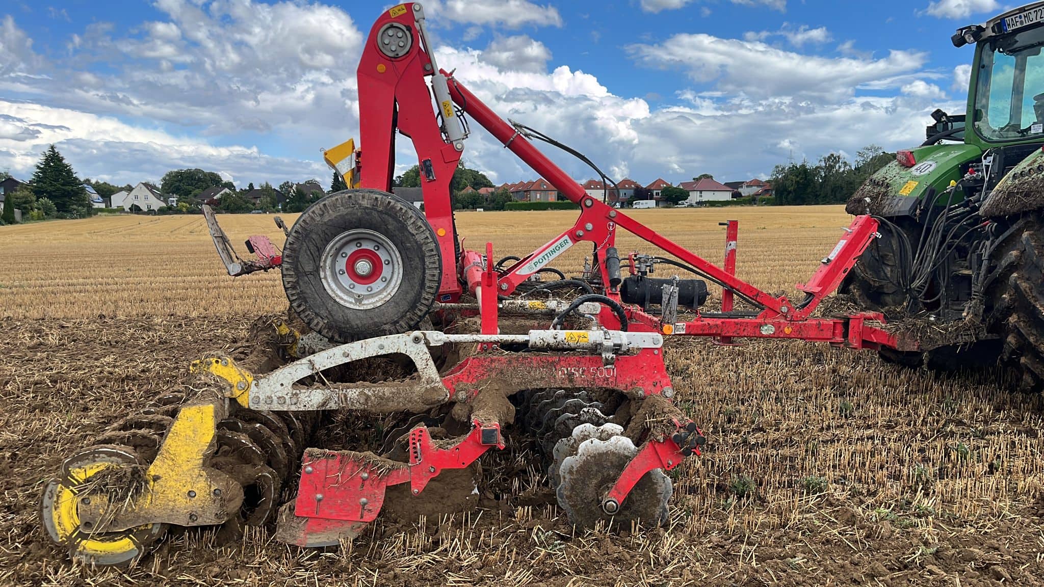 Eine rote Landmaschine pflügt ein Feld, daneben ein grüner Traktor unter blauem Himmel.